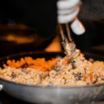 A close-up of creamy macaroni and cheese mixed with mushrooms being stirred in a skillet using tongs. The dish appears rich and savory, with steam rising gently.