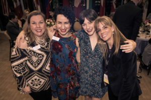Four smiling women stand closely together at an event, wearing a variety of dresses and name tags. They are in a warmly lit room with flags in the background and people seated at tables.