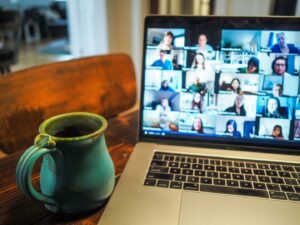 A laptop displaying a grid of people in an online video call is on a wooden table. Next to the laptop is a green ceramic mug filled with a dark beverage. The background is softly blurred, showing an interior setting.