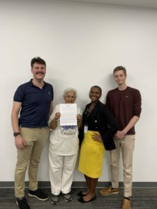 Four people stand in front of a white wall. An older woman in the center holds a paper, flanked by two men and a woman. They are smiling and casually dressed.