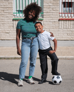 A smiling woman in a green striped shirt and jeans stands with her arm around a young boy in a white shirt and black pants. The boy stands with one foot on a soccer ball. They are outside on a paved area near a building.