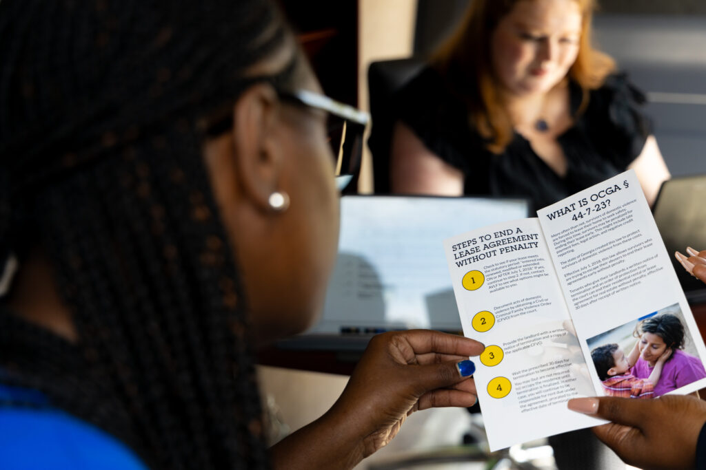 A woman in the foreground is reading a pamphlet with steps to end a lease agreement. In the background, another woman with red hair is focused on her laptop. The setting appears to be an office or meeting room.