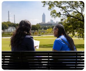 Two people sitting on a park bench with their backs to the camera. One is holding an open book. In the background, trees and a city skyline are visible under a clear sky.