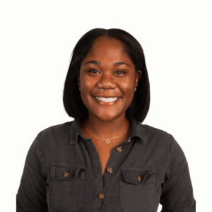 A woman with straight black hair smiles at the camera. She is wearing a dark button-up shirt and a gold necklace with a small pendant, standing against a plain white background.