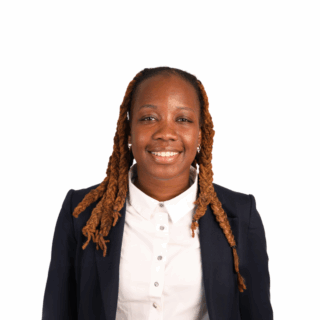 A woman with long, brown, twisted hair smiles at the camera. She is wearing a dark blazer over a white button-up shirt, standing against a plain white background.