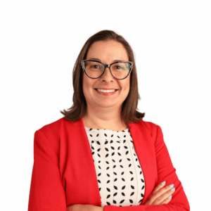 A woman with glasses, shoulder-length brown hair, and a big smile stands with arms crossed. She wears a red blazer over a white patterned blouse, posing against a plain white background.