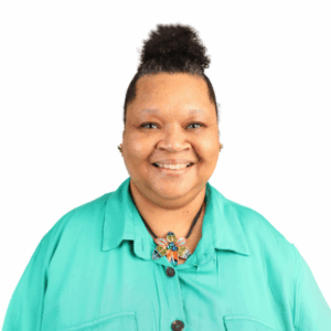 A woman with curly hair in a bun, wearing a turquoise shirt and a colorful beaded butterfly necklace, smiles at the camera against a white background.