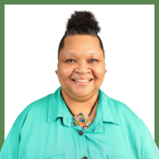 Smiling person with curly hair in a bun, wearing a turquoise shirt and a colorful butterfly-shaped necklace, standing against a white background.