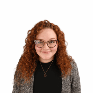 A person with curly red hair, wearing glasses, a grey blazer, and a black top, smiles while facing the camera against a plain white background.