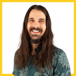 A person with long, dark hair and a beard is smiling at the camera. They are wearing a green shirt with a sunflower pattern. The background is white with a yellow border.