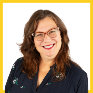 A woman with long, wavy brown hair wearing glasses and a navy shirt with floral embroidery smiles. The photo has a bright yellow border. The background is white.