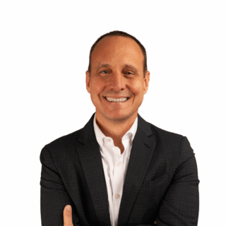 A man in a dark suit jacket and white shirt stands smiling with his arms crossed against a plain white background.