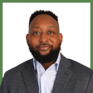 A man with a beard is wearing a blue shirt and a gray blazer, standing against a white background with a green border. He is smiling gently at the camera.
