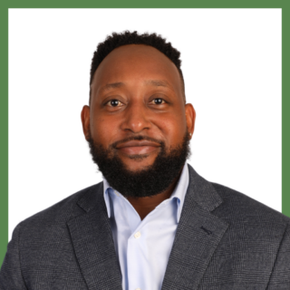 A man with a beard is wearing a blue shirt and a gray blazer, standing against a white background with a green border. He is smiling gently at the camera.