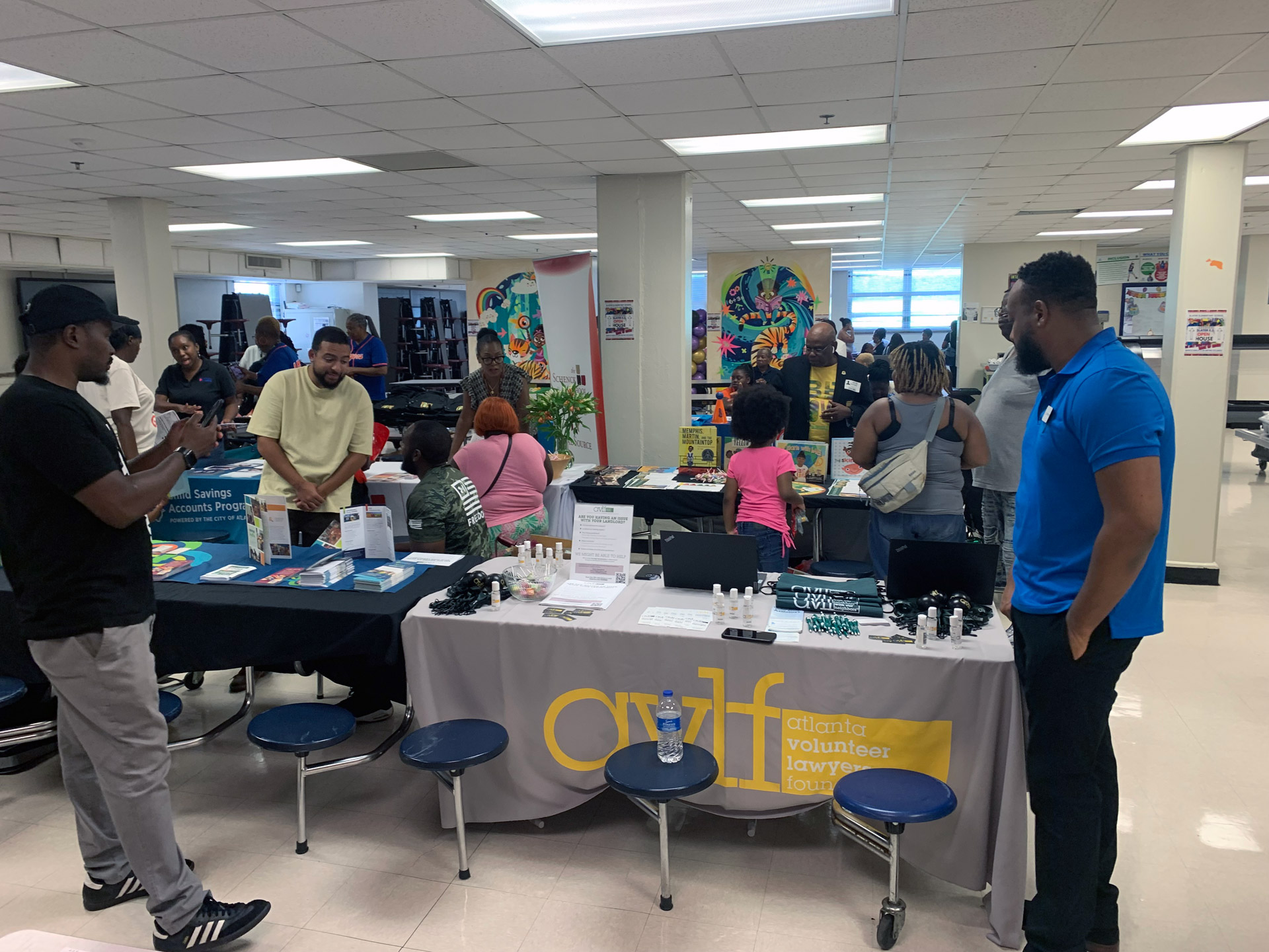 A community event is underway in a hall with several tables displaying information and materials. People are engaging and conversing. A table in the foreground features a banner for the Atlanta Volunteer Lawyers Foundation.