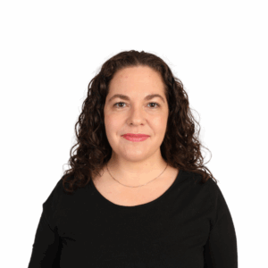 A woman with curly brown hair, wearing a black top and a thin necklace, smiles softly while facing the camera against a plain white background.
