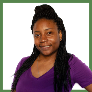 A person with long braided hair smiles at the camera, wearing a purple V-neck shirt and earrings. The background is plain white.