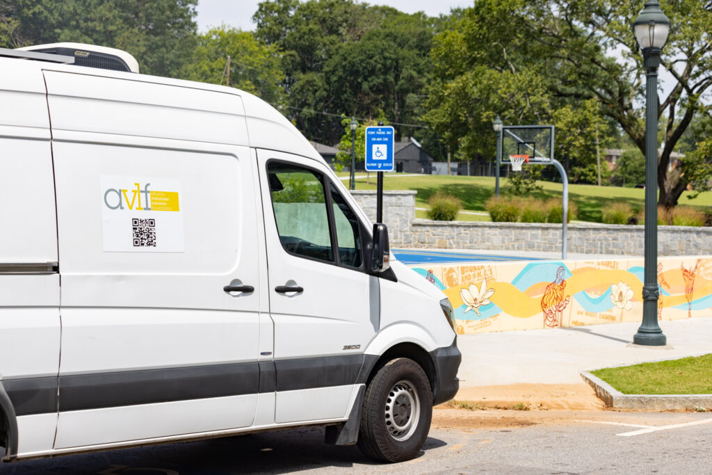 A white van with a company logo and QR code is parked beside a wheelchair-accessible parking space. In the background, there's a colorful mural on a low wall and a basketball court, with trees and a lamppost nearby.