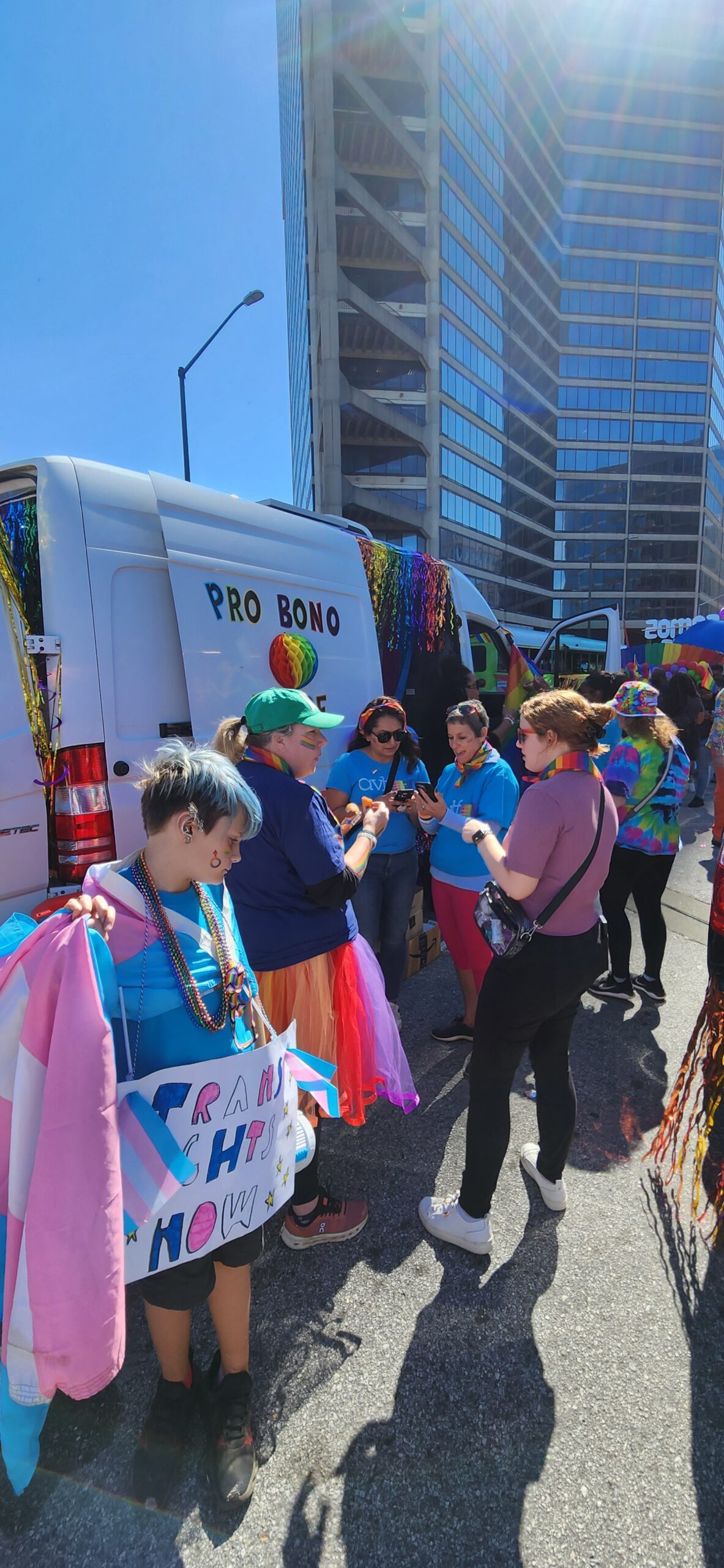 A group of people stand by a white van decorated with rainbow streamers at a street event. One person holds a βTrans Rights Now!β sign, and others wear colorful outfits and accessories. Tall buildings are visible in the background.