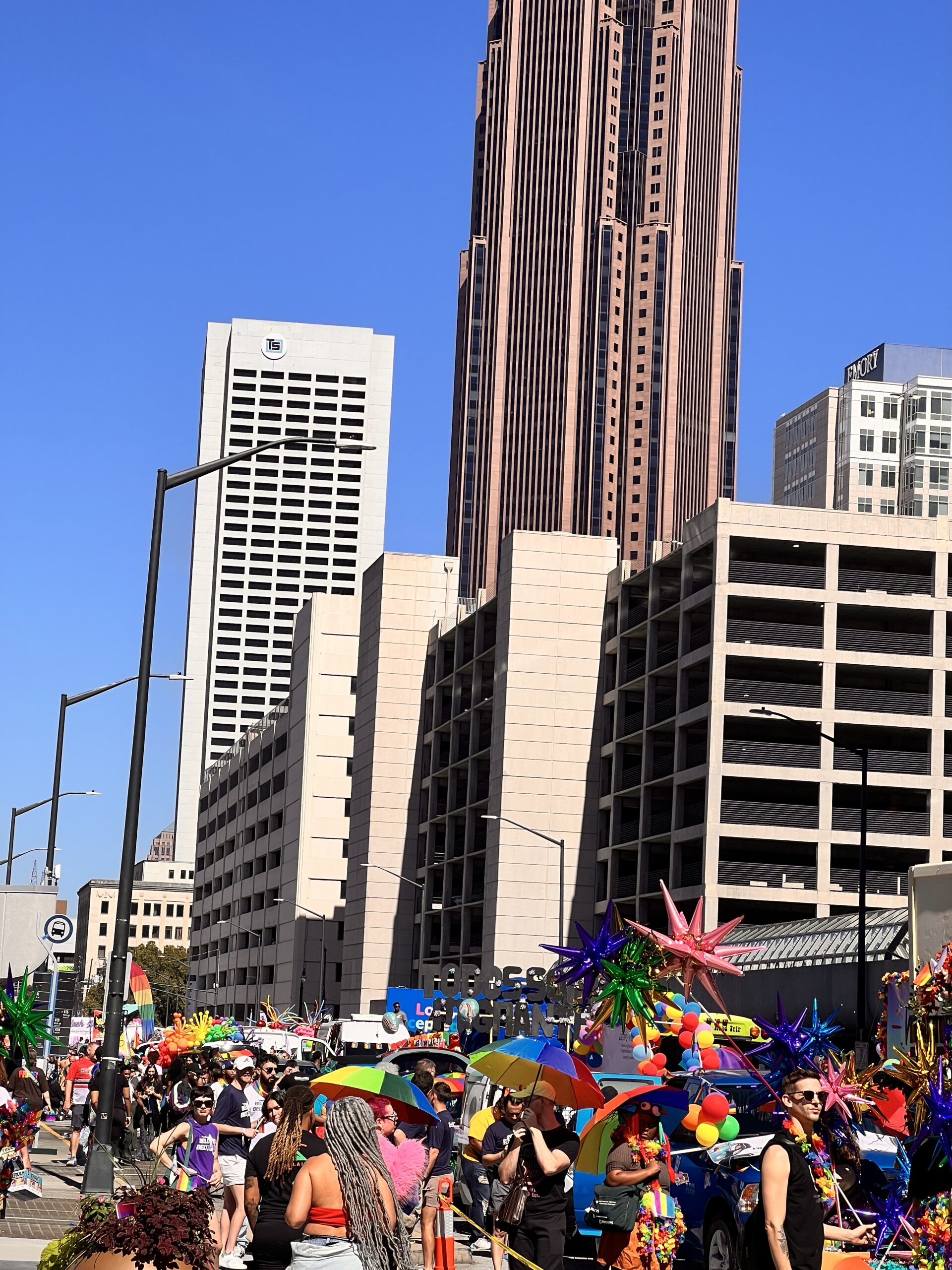 A lively parade with people in colorful outfits and rainbow decorations moves through a city street lined with tall buildings under a clear blue sky. Balloons and umbrellas add to the festive atmosphere.