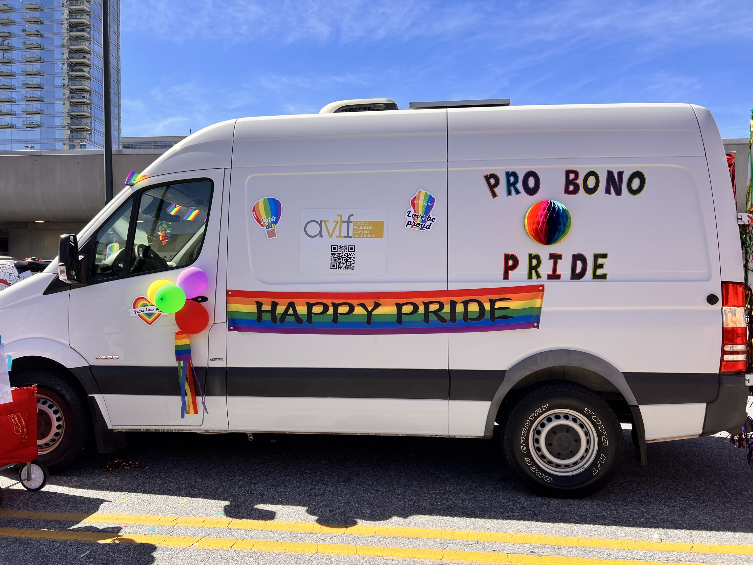 A white van decorated with rainbow balloons, a rainbow flag, and signs reading βPRO BONO PRIDEβ and βHAPPY PRIDE,β participates in a parade. The background shows a tall building and a clear blue sky.