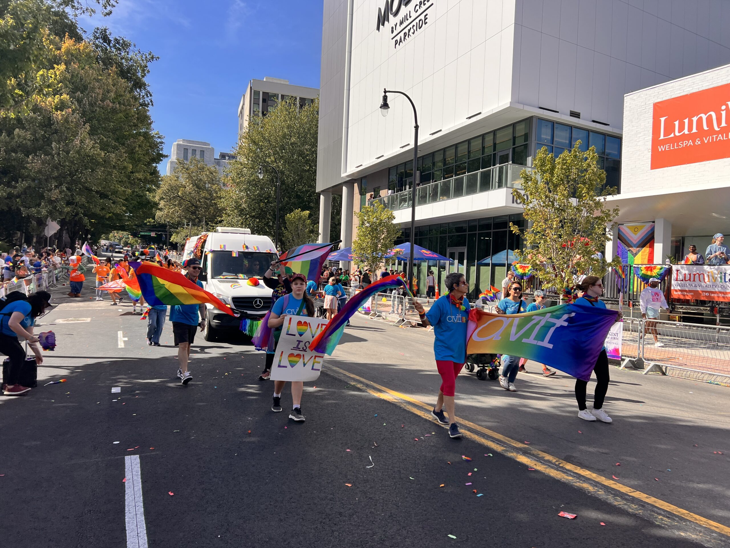 People march in a parade holding rainbow and pride flags, as well as signs reading βLoveβ and βAviv.β Spectators watch from sidewalks and a building. Itβs a sunny day with confetti scattered on the street.