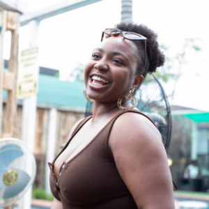 A woman with hoop earrings and sunglasses on her head smiles brightly outdoors. She is wearing a brown sleeveless top and standing in front of a fan, with blurred buildings and greenery in the background.