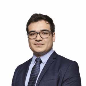 A man with short dark hair and glasses, wearing a navy suit, light blue shirt, and patterned tie, smiles softly while standing against a plain white background.