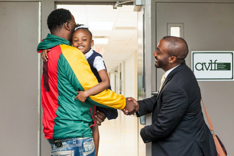 A man in a colorful jacket holds a smiling young girl as he shakes hands with another man in a suit at a doorway inside a building. The girl wears a school uniform and appears happy.