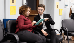 Two people sitting on chairs in an office setting, looking at a tablet together. One person points at the screen while the other holds the device. Yellow papers are posted on the wall behind them.