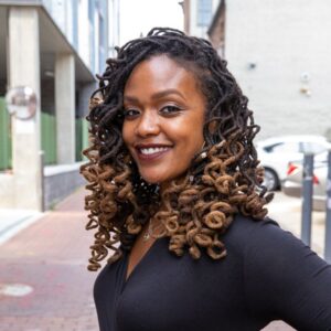 A woman with curly locs that are dark at the roots and lighter at the ends smiles confidently while standing outdoors on a city street, wearing a black top.