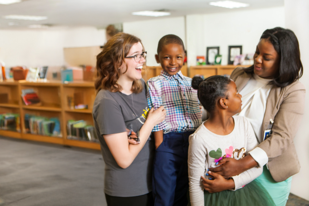 Two women and two children stand together in a library, smiling and talking. Bookshelves filled with books are visible in the background. The group appears happy and engaged.