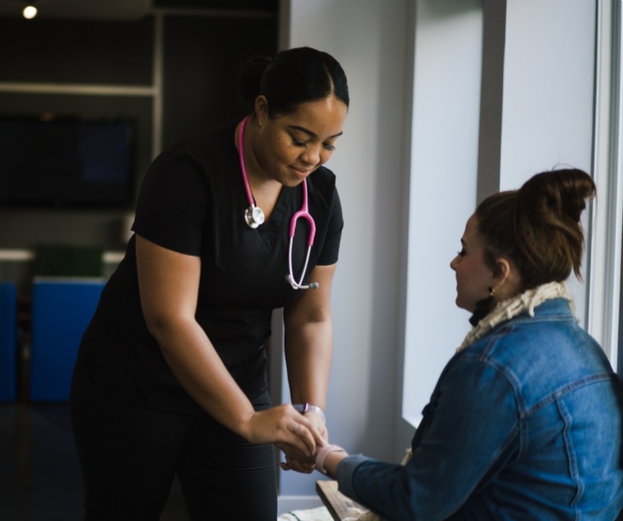 A healthcare worker with a stethoscope helps a seated woman by taking her pulse or checking her wrist in a brightly lit room near large windows.