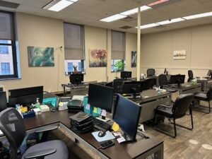 An empty office space with several desks, computers, office chairs, and monitors. Artwork decorates the walls, and natural light comes through windows with blinds. The room appears organized and tidy.
