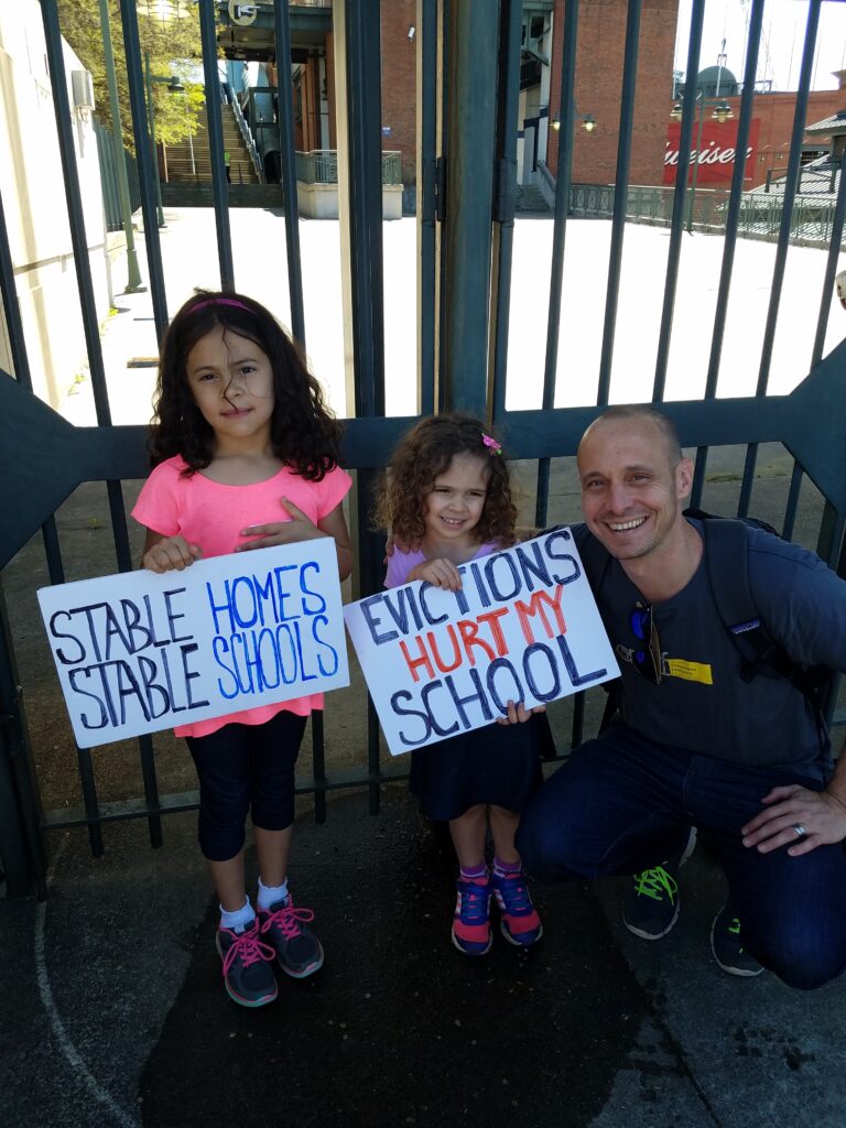 Two young girls hold signs reading "Stable Homes Stable Schools" and "Evictions Hurt My School" while standing next to a smiling man crouched beside them. They are outdoors in front of a metal gate.