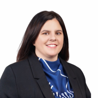 A woman with straight dark brown hair, wearing a black blazer and a blue patterned scarf, is smiling against a plain white background.