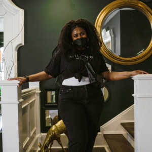 A woman with curly hair stands between two white banisters in a stylish room with dark walls, circular mirrors, and a decorative gold animal figure visible behind her. She is wearing a black t-shirt and jeans.