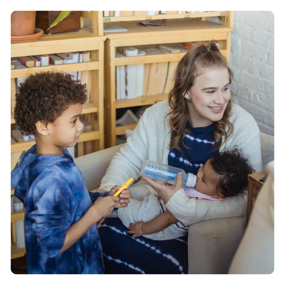 A woman sits in a chair smiling while bottle-feeding a baby in her arms. A young boy stands nearby holding a yellow toy, with shelves of books and plants in the background.