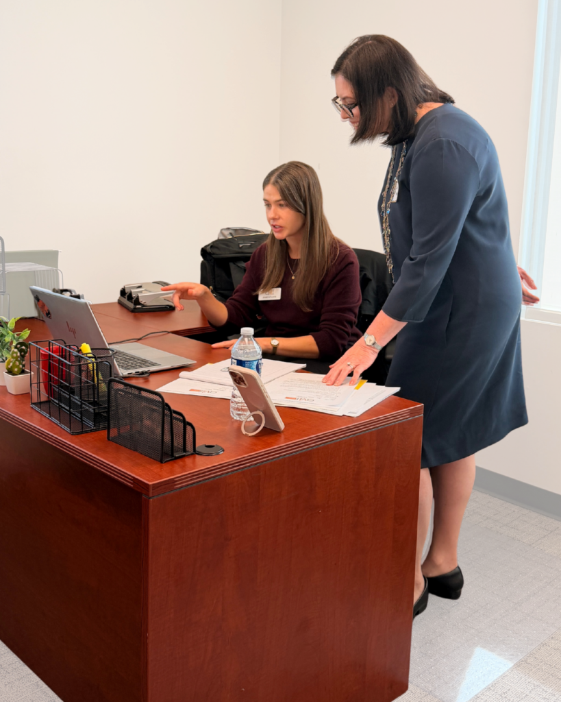 Two women in an office are working together at a desk. One is seated, looking at a laptop and pointing at the screen, while the other stands beside her holding papers. The desk has office supplies, water bottles, and documents on it.