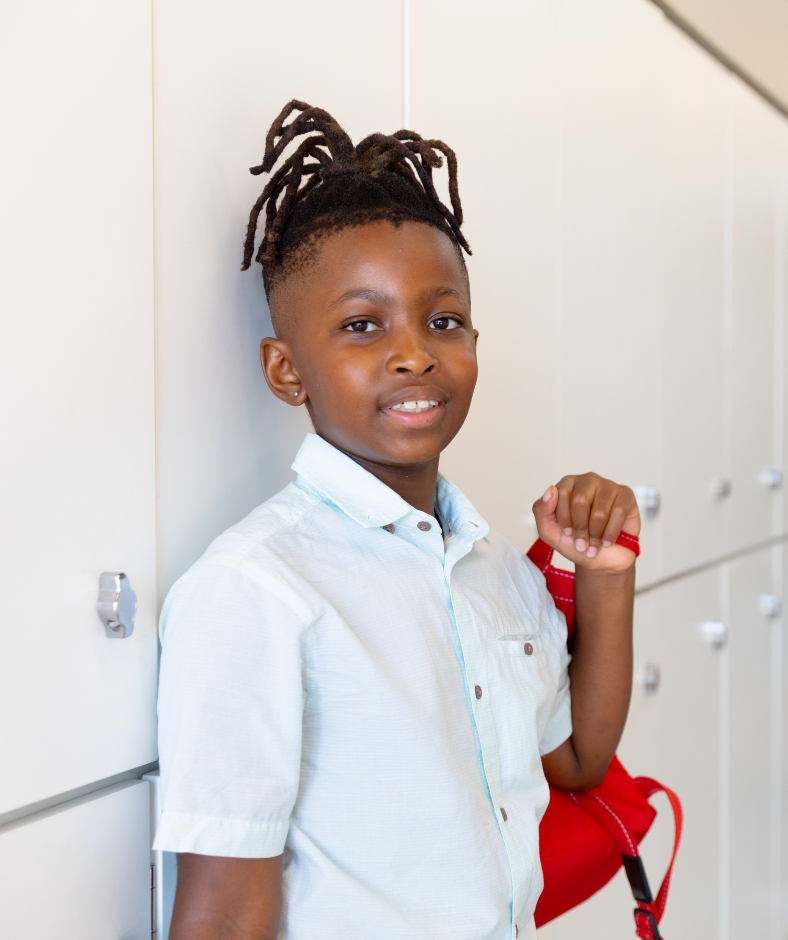 A young boy with short dreadlocks stands smiling by lockers, wearing a light blue shirt and holding a red backpack over his shoulder.