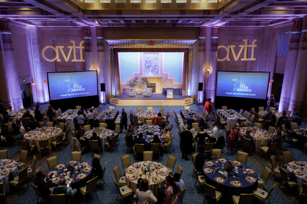 Large ballroom set up for an awards event, with round tables filled with guests, two large screens displaying “AVLF” and “5th Annual AVLF Awards,” and a stage with a podium and floral decorations at the front.