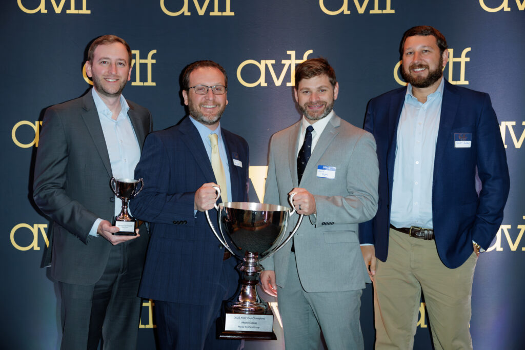 Four men in business attire stand smiling in front of a backdrop with "avjf" logos, holding two large trophies, suggesting a celebratory or award event.