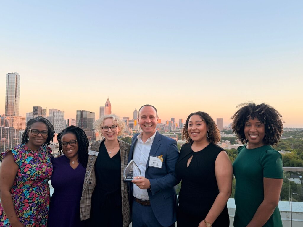 Six people stand together smiling on a rooftop with a city skyline at sunset in the background. The person in the center holds a glass award.