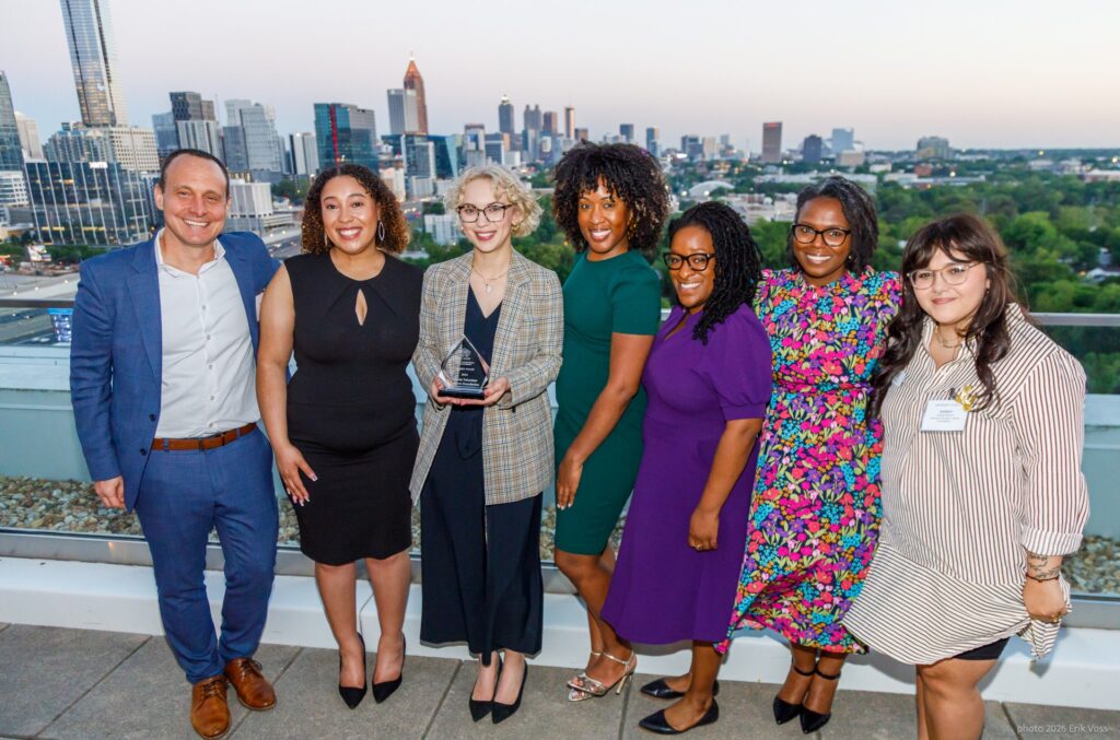 Seven people stand smiling on a rooftop with a city skyline in the background. The group includes six women and one man; one woman holds an award. It appears to be a celebratory or professional event at sunset.