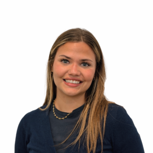 A woman with long, straight light brown hair is smiling at the camera. She is wearing a navy blue top, a black sheer undershirt, and a gold chain necklace. The background is plain white.