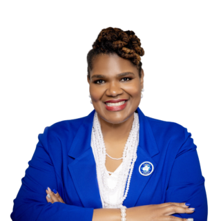 A woman wearing a royal blue blazer, white top, and pearl jewelry smiles confidently with her arms crossed against a white background. She has styled hair and wears a blue pin on her lapel.