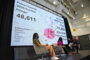 Three people sit on stage in front of a large screen displaying eviction data for Fulton County, including a map and statistics such as “2025 Eviction Filings: 48,611” and “Six-Cluster Filings: 11,955 (25%).”.