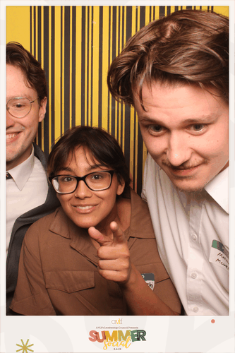 Three people pose in a photo booth with a yellow striped backdrop. One person in the center, wearing glasses, points towards the camera, while the others smile. The frame reads "SUMMER Social.