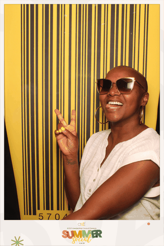 A smiling woman wearing sunglasses and a white top flashes a peace sign in front of a large yellow and black barcode backdrop. The photo has a "SUMMER SZN" logo at the bottom.
