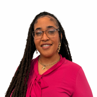 A woman with long, braided hair wears glasses, gold jewelry, and a bright pink top with a bow. She is smiling and standing against a plain white background.